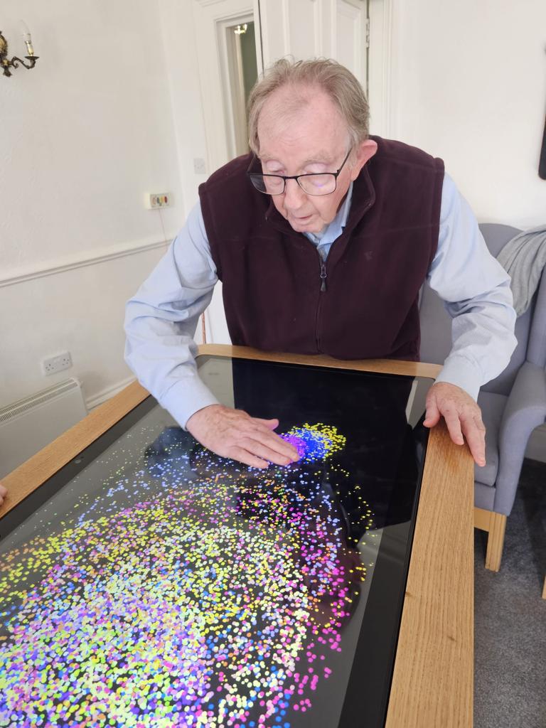 Elderly man engaging with colorful sensory sand on a glass table, promoting tactile interaction and creativity in a cozy indoor setting.