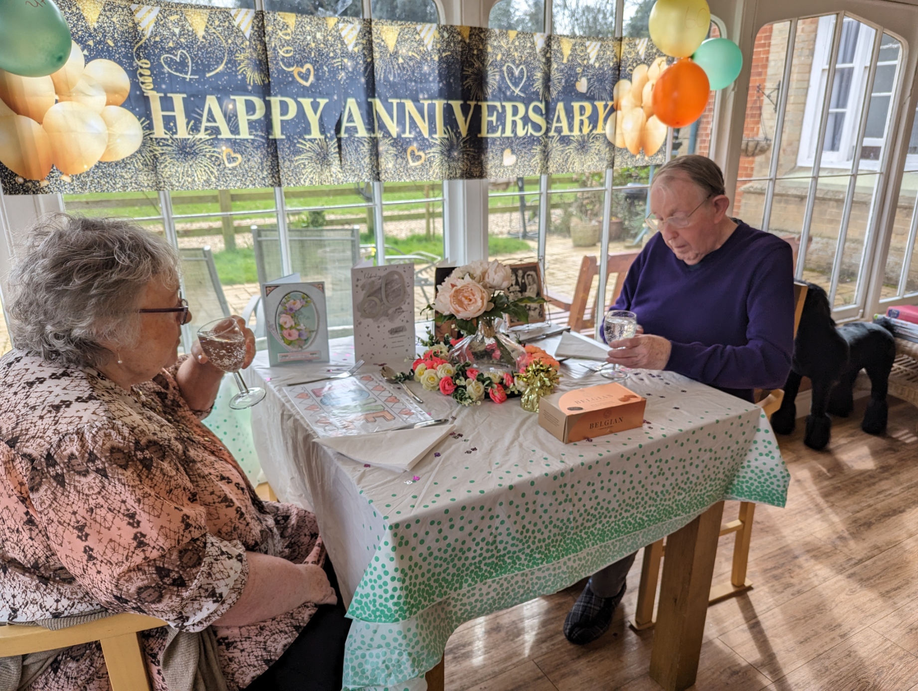 Elderly couple celebrating their anniversary at a decorated table with flowers, cards, and balloons, enjoying a cozy indoor setting.