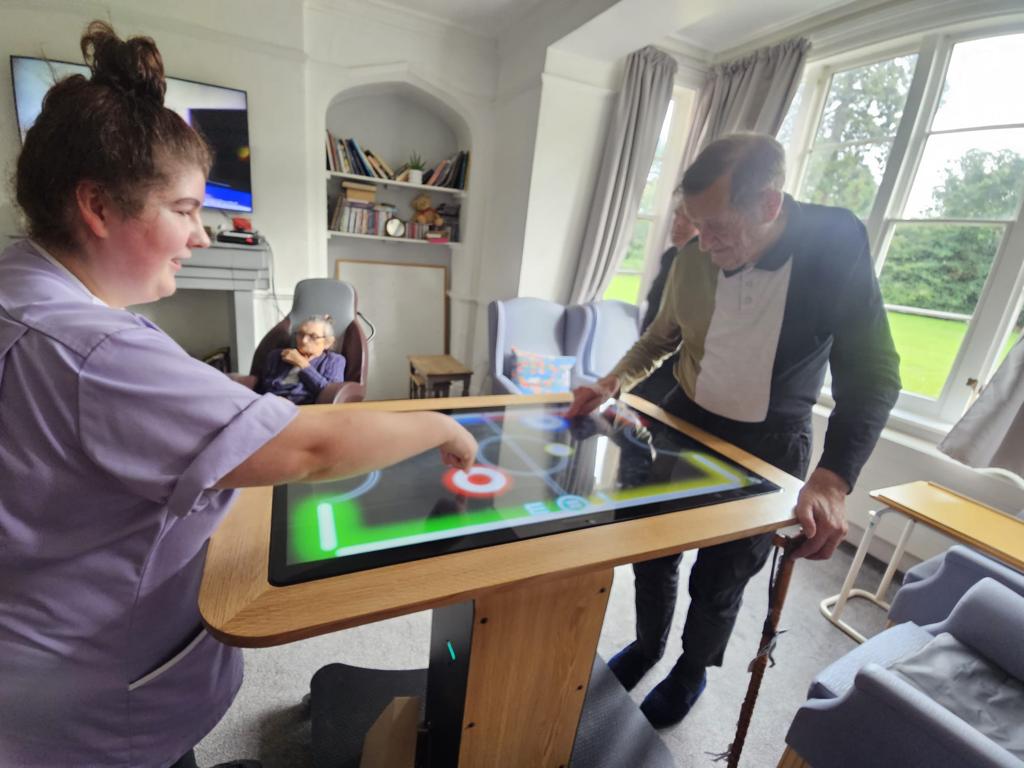 Interactive touchscreen table being used by a woman in a purple shirt and a man in formal attire, engaging in a game or activity, with two people observing in a cozy indoor setting with natural light.