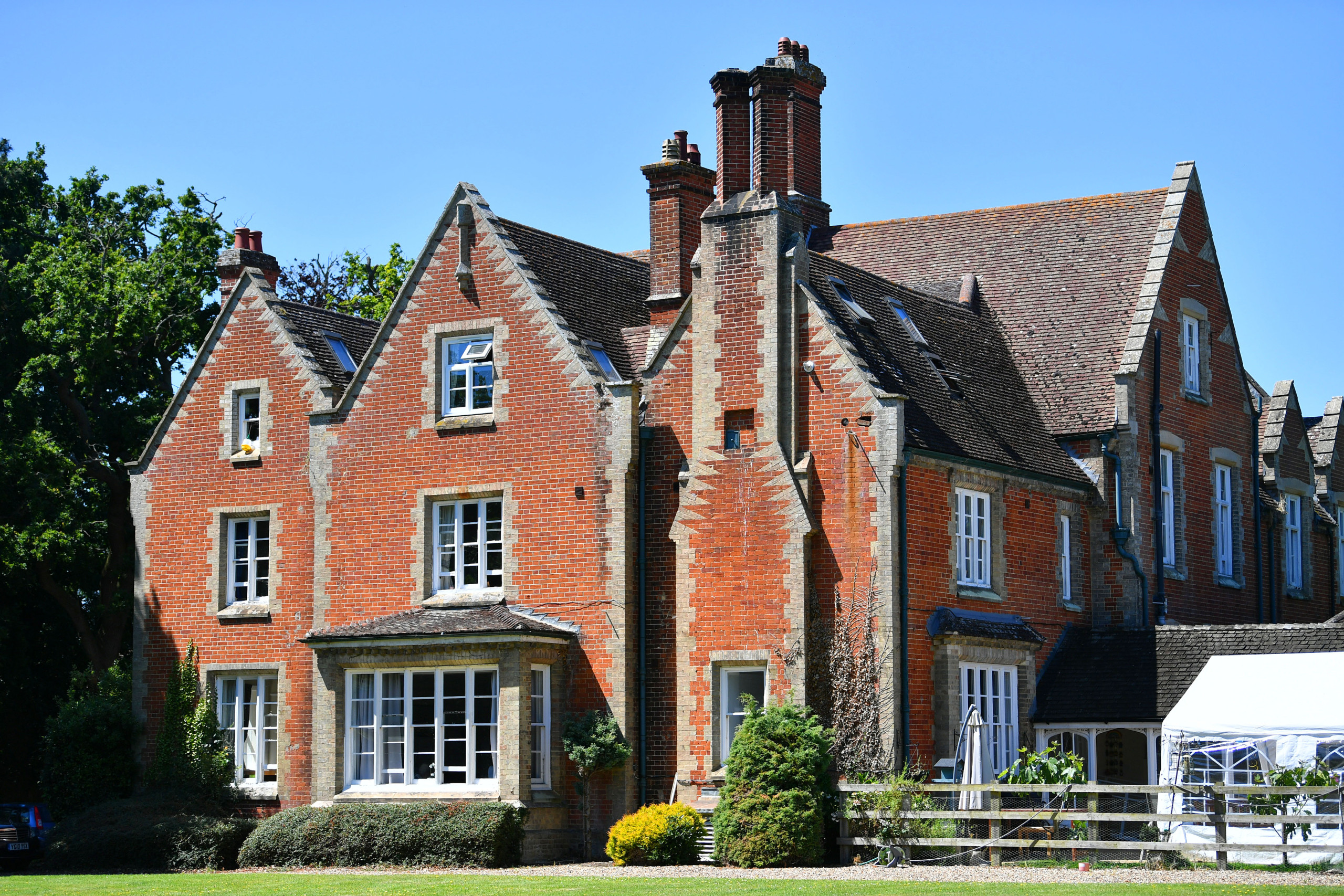 Historic red brick house with multiple chimneys and large windows surrounded by greenery on a clear sunny day.