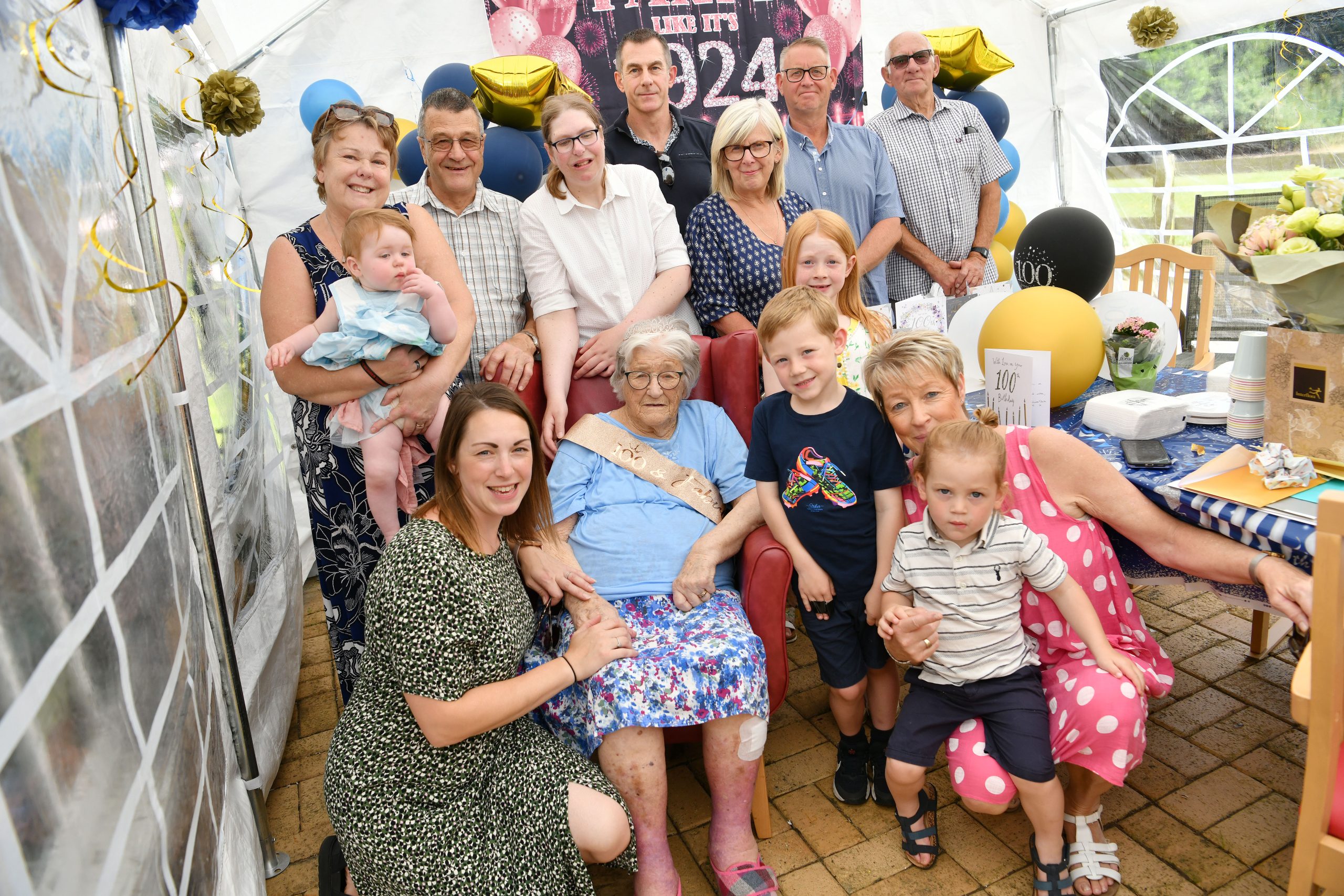 Family gathering celebrating a milestone birthday with a large group of people, including children and adults, in a decorated tent with balloons and a banner.
