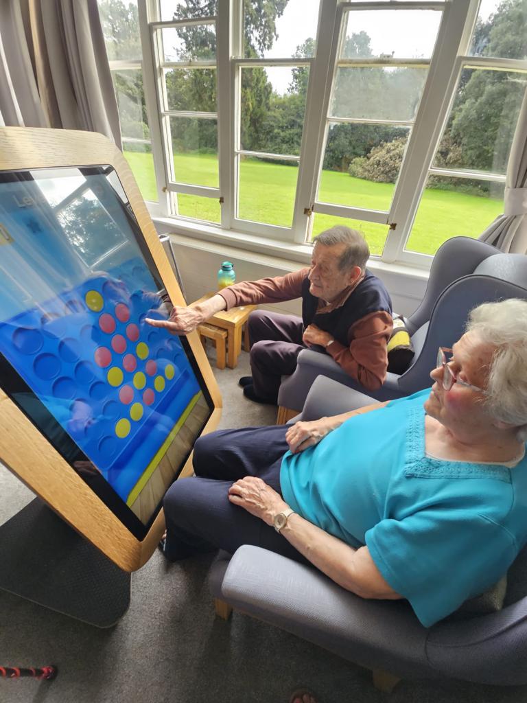 Elderly individuals engaging with a large interactive touchscreen game in a bright, airy room with large windows, promoting cognitive activity and social interaction.