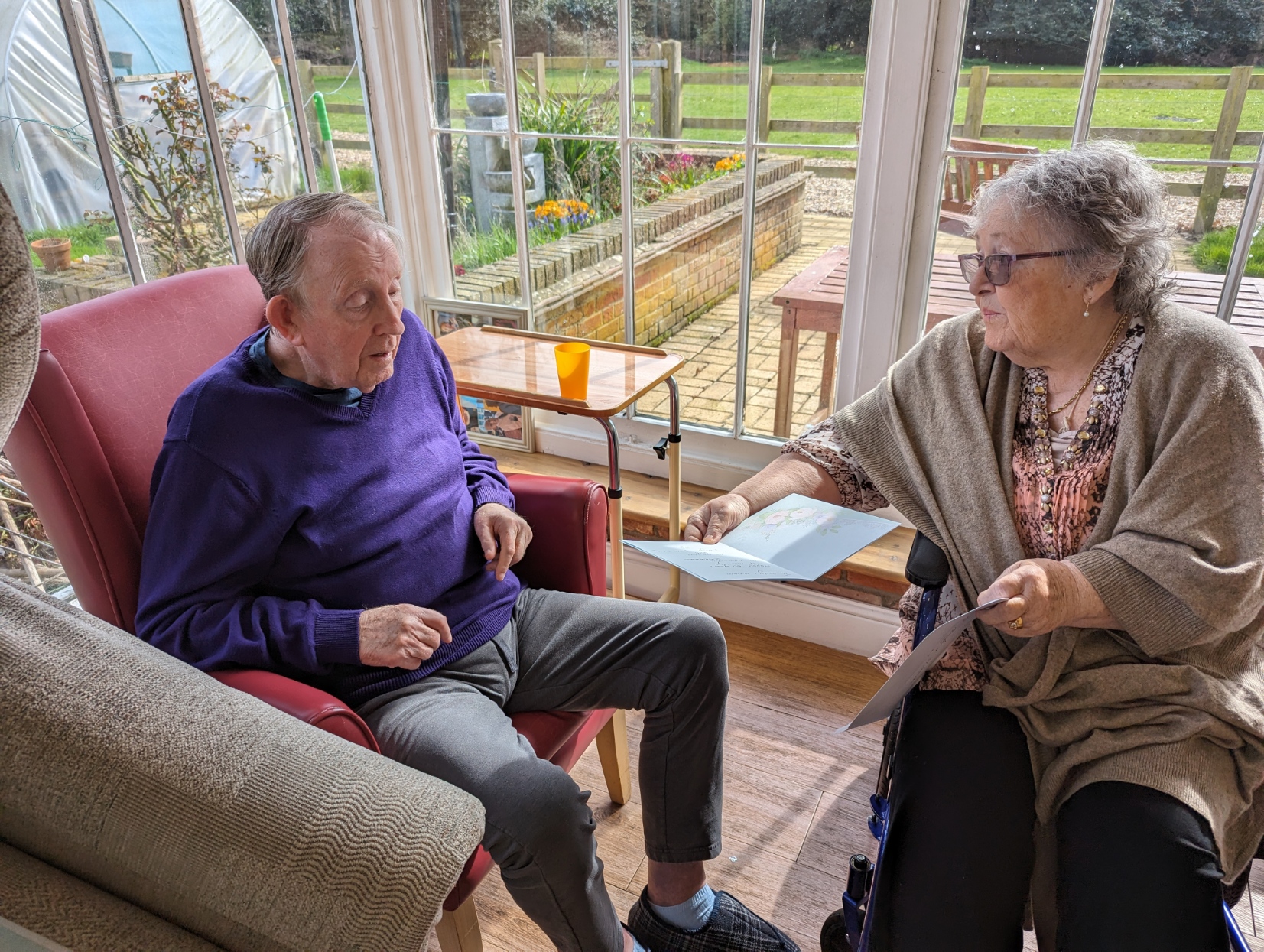 A man in a purple sweater and a woman in a shawl engage in conversation while sitting in a sunlit room, surrounded by greenery and outdoor seating.