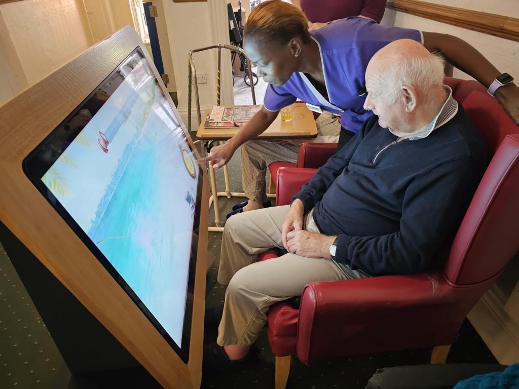 A caregiver assisting an elderly man with a touchscreen interactive display in a comfortable setting, promoting engagement and cognitive activity for seniors.