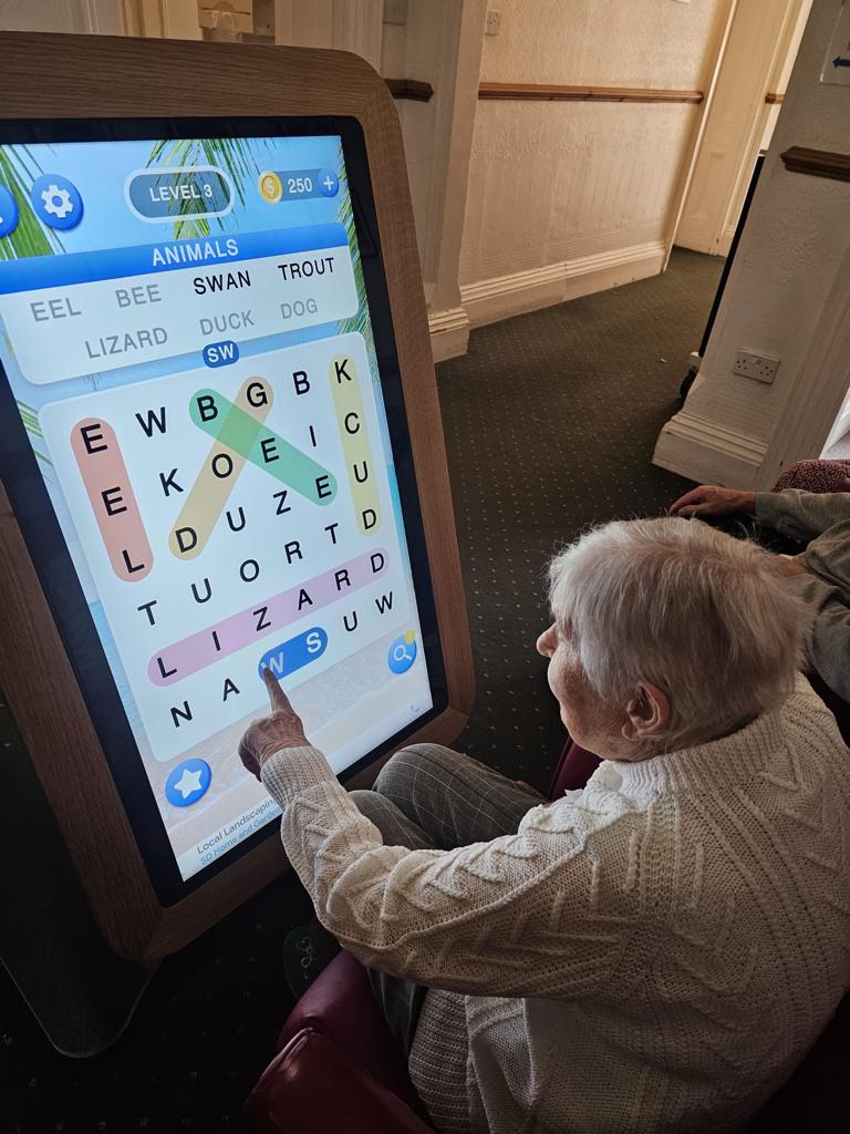 Senior woman interacting with a large touchscreen display while playing a word game, focusing on finding the word "lizard" among other letters.