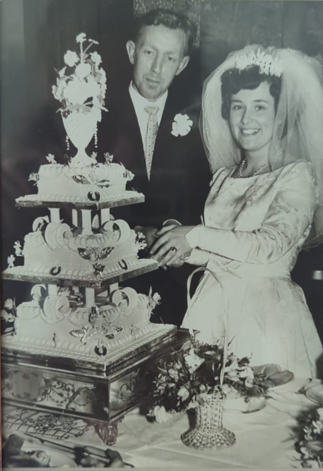 Black and white photo of a smiling bride and groom standing beside an elaborate wedding cake, showcasing a classic wedding celebration. The bride is wearing a lace gown with a veil, while the groom is in a tuxedo, both looking joyful at their special moment.